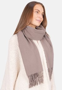 Femme aux longs cheveux bruns portant un pull en tricot blanc et une écharpe taupe à franges enroulée autour du cou, regardant sur le côté.