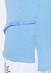 Světle modré tričko s texturovaným povrchem, které má třepený detail podél bočního švu a šikmý lem pro přidanou eleganci.