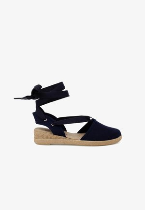 Espadrille de color azul marino con parte superior de lona, suela de yute trenzado y tirantes de tela entrelazados. Presenta un diseño de puntera cerrada y forma redondeada.