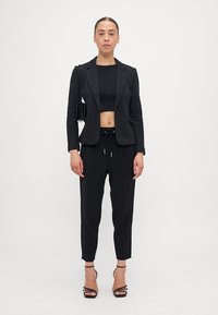 Črn prirezani blazer nad črnimi krojenimi topom, skupaj s črnimi hlačami na vrvico in črnimi sandali z naramnicami. Gladek material, minimalistična zasnova.