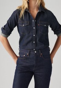 Mörk denimskjorta med knappar och två bröstfickor, kontrasterande sömmar och uppvikta ärmar; ihop med högmidjade mörka denimjeans.