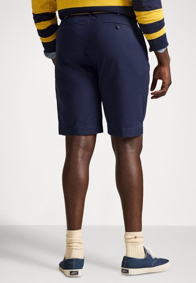 Tengerészkék rövidnadrág két hátsó zsebbel, szabott kialakítással és felhajtott szegéllyel, kék cipővel és krémszínű zoknival párosítva.