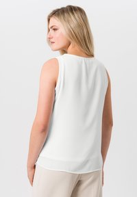 Haut sans manches blanc avec un décolleté rond, tissu lisse, détail de plis au dos et ourlet incurvé, associé à un pantalon beige.