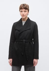 Μαύρο παλτό με γιακά σαρίλ, ζώνη για δέσιμο, μακριά μανίκια και απαλή υφή. Φορεμένο πάνω από ριγέ πουκάμισο, παρουσιάζεται μπροστά από απλό φόν το.