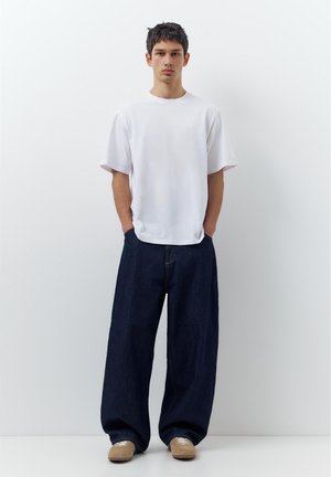 Fiatal férfi fehér sima pólóban, széles sötétkék farmernadrágban és világosbarna tornacipőben, kezeit a zsebében tartva, egyszínű fehér háttér előtt áll.