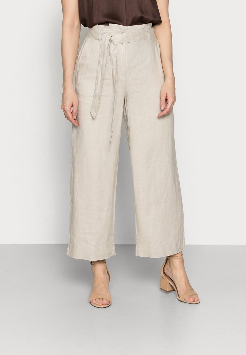 Pantalon large en lin beige avec taille ceinturée et poches latérales. Texture lisse, ourlet longueur cheville, associé à des sandales à talons hauts nude.