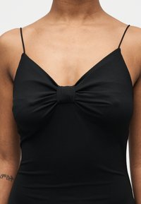 Personne portant un haut noir sans manches avec des fines bretelles et un détail de nœud froncé au centre de la poitrine.