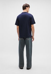 Ναυτικό μπλε βαμβακερό μπλουζάκι με στρογγυλή λαιμόκοψη, κοντά μανίκια και άνετη εφαρμογή. Φοριέται με χαλαρά ανοιχτό μπλε τζιν παντελόνια. Όψη από πίσω.