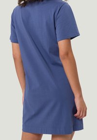 Personne portant une robe bleue à manches courtes et à mi-longueur, se tenant de dos face à un fond uni.