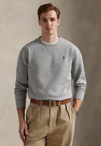 Jonge man draagt een grijze crewneck trui met blauw logo, beige geplooide broek, bruine riem en zilveren horloge, staand voor een effen achtergrond.
