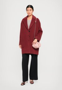 Manteau en mélange de laine bordeaux avec de grands revers, deux poches avant et trois boutons noirs. Associé à un pantalon noir à jambes larges. Sac à main rose.