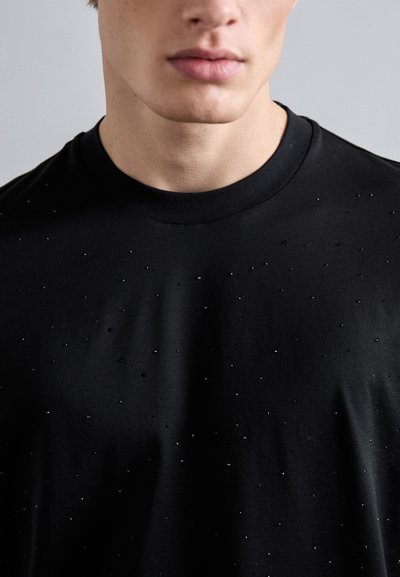 Svart T-skjorte med rund hals og små, diskrete pynting på foran, laget av mykt bomullsstoff. Jevn tekstur.