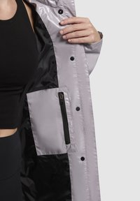 Jachetă impermeabilă de culoare mov deschis, cu o textură lucioasă, având un căptușeală neagră și un buzunar interior cu fermoar. Se închide cu capse pe față.