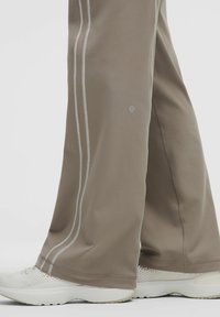 Világosbarna sportnadrág sima anyagból, két függőleges fehércsíkkal az oldalán és egy logóval az alján.