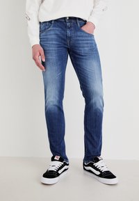 Man som bär slim-fit blekt blå jeans och svarta sneakers med vita snören, står med en hand i fickan mot en enfärgad bakgrund.