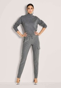 Vrouw poseert zelfverzekerd in een grijze coltrui, grijze slim fit cargobroek met trekkoord en zilveren spitse hakken.