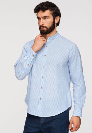 Homme aux cheveux bouclés foncés et à la barbe, portant une chemise à manches longues rayée bleu clair et blanc avec des boutons en bois, posant avec la main près du menton.