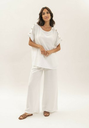 Femme debout portant un haut blanc ample avec des boutons décoratifs sur les manches, un pantalon large blanc et des sandales plates marron sur fond neutre.