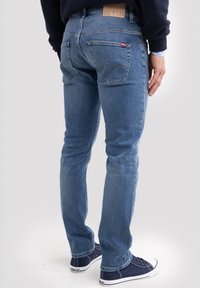 Vyras dėvi šviesiai mėlynas prigludusias džinsines kelnes ir tamsiai mėlynas sportbačius, stovėdamas viena ranka laisvai nuleidęs prie šono.