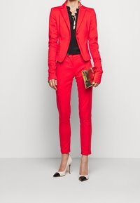 Czerwony marynarka dopasowana i odpowiadające jej wąskie spodnie, połączone z czarną bluzką i metaliczną torebką. Buty mają szpiczasty nos z kontrastującymi akcentami.