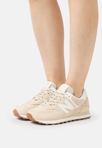 Beige semišové tenisky s bílými akcenty, vybavené polstrovaným límcem, šněrováním a gumovou podrážkou. Číslo modelu 574 viditelné na jazyku.