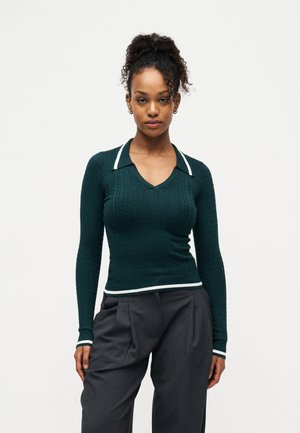 Žena s kučeravými vlasmi v tmavozelenej pletenej blúzke s bielym lemom a tmavosivými plisovanými nohavicami, stojaca na jednofarebnom bielom pozadí.