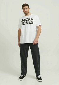 Giovane in piedi che indossa una maglietta bianca "JACK & JONES", pantaloni neri e sneaker nere a basso profilo con suole e lacci bianchi.