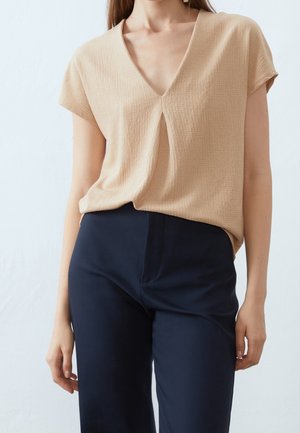 Vrouw die een beige, kort mouwens, gestructureerd V-hals top draagt met een plooivorm aan de voorkant en donkerblauwe, op maat gemaakte pantalon, staand met ontspannen armen.