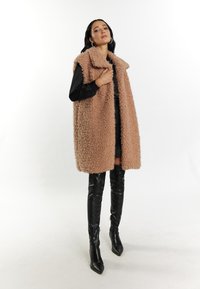 Übergro west, flauschig in Beige mit einem Kragen. Getragen über einem schwarzen Oberteil und einem Minirock. Kombiniert mit hohen schwarzen Stiefeln mit spitzer Zehenform.
