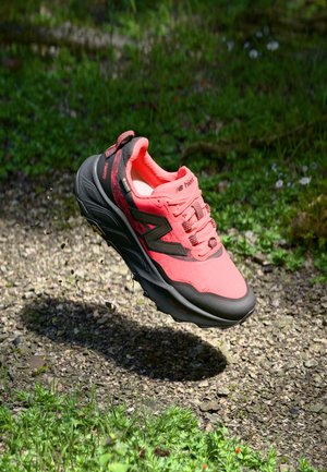 Bright pink and black New Balance running shoe suspended mid-air above a gravel path with green grass background.