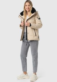 Beige puffer jacket with a black fur-lined hood, zip and snap closures, paired with gray jeans and white sneakers. Textured material.