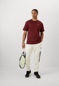 Burgunderfarget bomull T-skjorte med en liten logo, sammen med hvite, slim-fit bukser med grå detaljer. Modellen holder et tennisracket.
