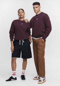 Dois modelos vestem roupas da Nike: um com um sweatshirt bordô e calções pretos, o outro com um sweatshirt bordô combinado com calças cargo castanhas, ambos com ténis.