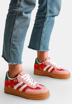 Chaussures en tissu floral rouge, détails en suède rose, rayures blanches, accents verts sur le talon, et semelles épaisses marron portées avec un jean bleu à ourlet effiloché.