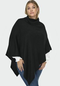 Poncho en tricot noir avec un col roulé haut, présentant une finition texturée et un design ample et drapé. Porté sur une chemise blanche et un jean bleu.