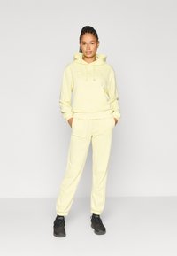 Ελαφρώς κίτρινο φούτερ με κορδόνια και λογότυπο, συνδυασμένο με αντίστοιχο παντελόνι φόρμας. Και τα δύο έχουν απαλή υφή και άνετη εφαρμογή. Μαύρα παπούτσια ολοκληρώνουν την εμφάνιση.