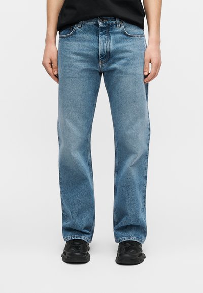 Personne portant un jean droit bleu clair, des baskets noires et un haut noir, debout devant un fond blanc uni.