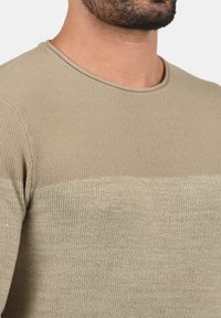 Pull beige en tricot avec une texture côtelée, présentant un col rond et un motif dégradé subtil dans des tons beige clair et foncé.