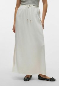 Jupe maxi blanche avec taille à cordon de serrage, dot�ée de deux fentes sur les côtés ; texture lisse ; associée à des chaussures plates noires.