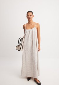 Šviesiai kreminė maksi suknelė su plonomis spaghetti petnešomis, įrubu paviršiumi ir laisvu siluetu. Modelis laiko švelnų kreminės spalvos rankinį krepšį su juodais akcentais.