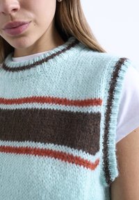 Κοντινό πλάνο γυναίκας που φοράει ανοιχτό μπλε πλεκτό γιλέκο με καφέ και πορτοκαλί οριζόντιες ρίγες πάνω από λευκό κοντομάνικο πουκάμισο.