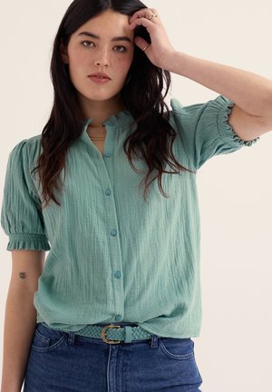 Donna con lunghi capelli scuri che indossa una camicetta verde chiaro con bottoni e cintura intrecciata, si tocca i capelli con la mano sinistra.