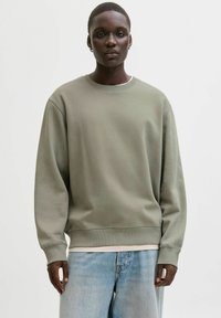 Olivzaļš džemperis ar apaļu apkakli un garām piedurknēm, ar ribveida aprocēm un apakšmalu. Valkāts virs gaiši krāsotas krekla kārtas un kombinēts ar gaišām džinsa biksēm.