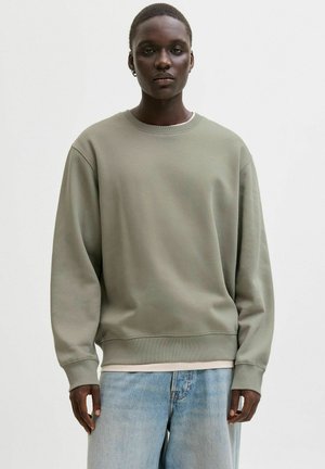 Olijfgroene trui met ronde hals en lange mouwen, voorzien van een geribbelde boord aan de mouwen en zoom. Gedragen over een lichte laag en gecombineerd met lichte jeans.