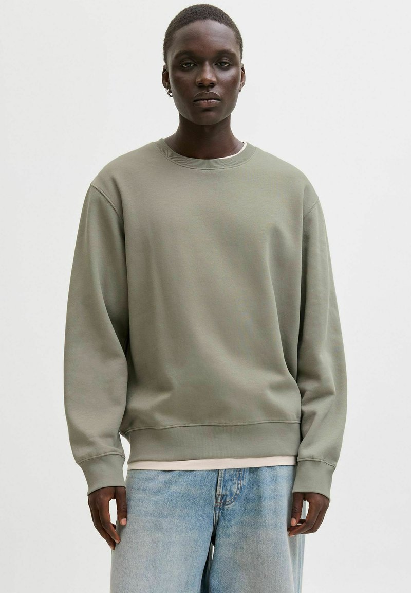 Olivzaļš džemperis ar apaļu apkakli un garām piedurknēm, ar ribveida aprocēm un apakšmalu. Valkāts virs gaiši krāsotas krekla kārtas un kombinēts ar gaišām džinsa biksēm.