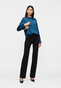 Γυναίκα που φοράει μπλούζα μακρυμάνικη μπλε γυαλιστερή, μαύρο παντελόνι, μαύρες γόβες, κρατώντας μια μικρή ασημί τσάντα στον ώμο της, στέκεται μπροστά σε λευκό φόντο.