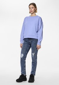 Lilac sweatshirt with a round neckline, loose fit, and ribbed cuffs paired with distressed blue jeans and black sneakers.