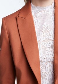 Blazer color ruggine con rever affilati, abbinato a un top bianco in pizzo con motivi floreali. Il materiale è liscio e strutturato.