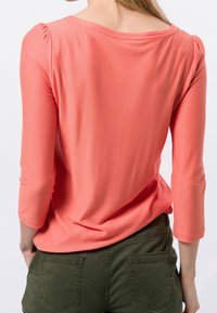 Top lungo coral dalle texture con scollo ampio e maniche a sbuffo, abbinato a pantaloni verde oliva con tasche posteriori.