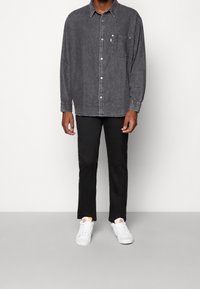 Camicia in denim grigia con maniche lunghe e taschino sul petto, abbinata a pantaloni neri slim e sneakers bianche. Texture liscia, design casual.